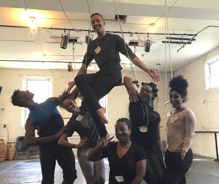 Six smiling people gathered around 1 person sitting in low trapeze, all dressed in dance clothing