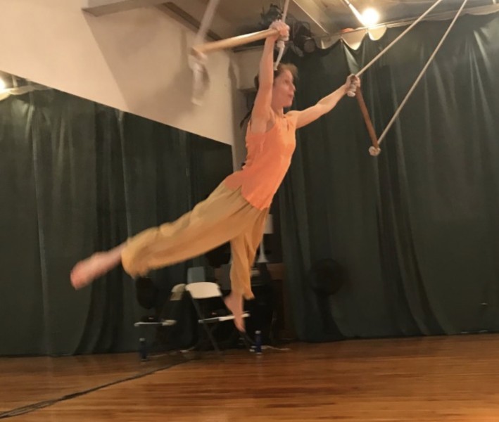 Woman with arms and legs out stretched while flying by hanging on to two trapeze bars, wearing orange shirt and flowing pants