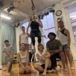 Six smiling people gathered around 1 person sitting in low trapeze, all dressed in dance clothing