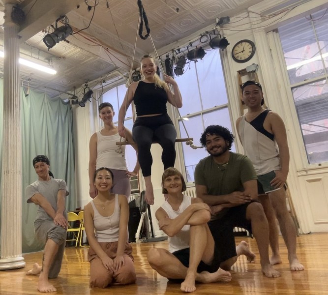 Six smiling people gathered around 1 person sitting in low trapeze, all dressed in dance clothing