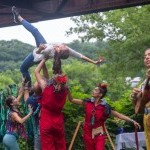 A dancer is held up by 4 other dancers in the air, a musician sings, all are in colorful costumes performing outside