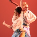 Two dancers in cream tops and blue and mustard skirts with orange background