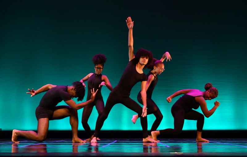Teen dancers in dark unitards and bare feet dance on a stage