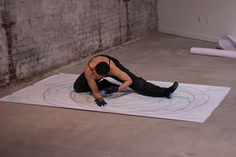 A dancer in black clothing dances on the floor on a large piece of paper.