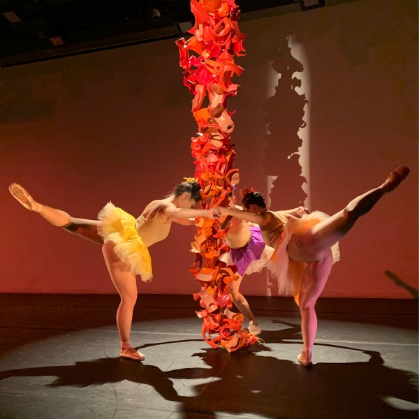 Three dancers with leg extended, surround a hanging orange sculptural column
