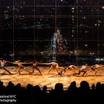 Ten contemporary dancers reach across a large stage, holding hands, with glass windows looking over Columbus Circle in NYC