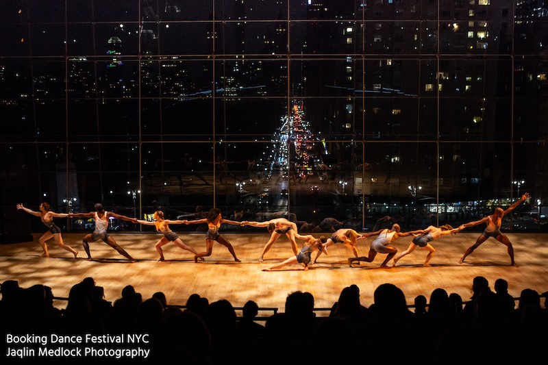 Ten contemporary dancers reach across a large stage, holding hands, with glass windows looking over Columbus Circle in NYC