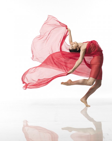 Dancer leans forward and balances on one leg with shear, red dress flowing behind her