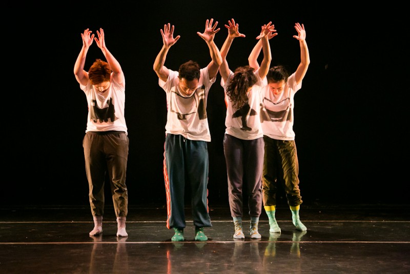 Four dancers stand with both hands raised in the air, palms facing forward, and head bowed down