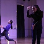 In a purple-hued lit stage space are two women with bent elbows, one with black hair and a blue shirt in profile with bent knees