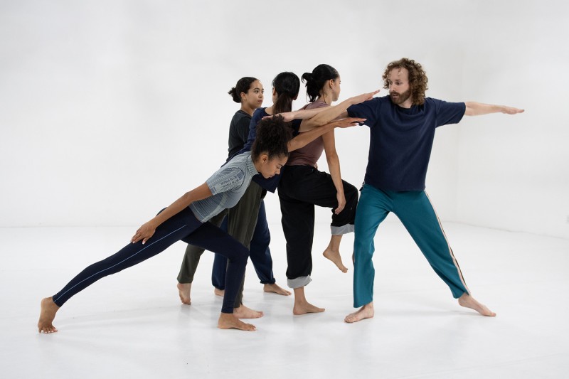 Five dancers are close together creating a cluster in a white dance studio.