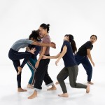 Five dancers are close together creating a cluster in a white dance studio.