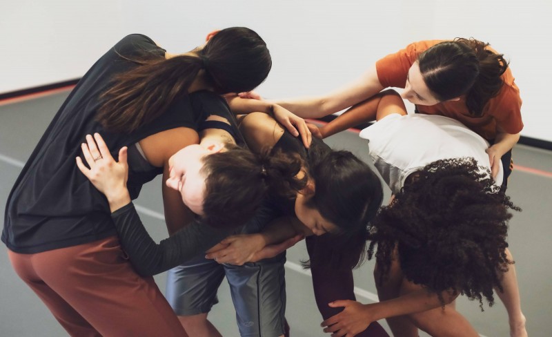 Five dancers create a sculptural shape. Close together, their arms overlap with knees bent, faces turned away from the camera.