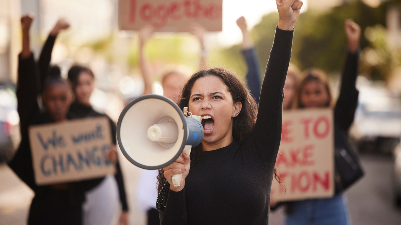 A stock image of people protesting. One person stands in the foreground, shouting into a megaphone. People behind them hold cardboard DIY signs