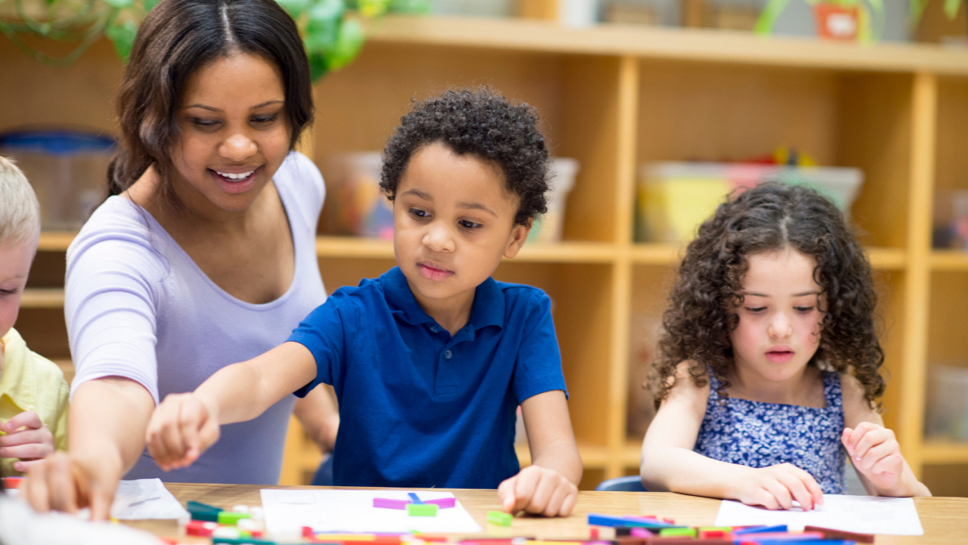 Children in a classroom, coloring, with a teacher behind them assisting.